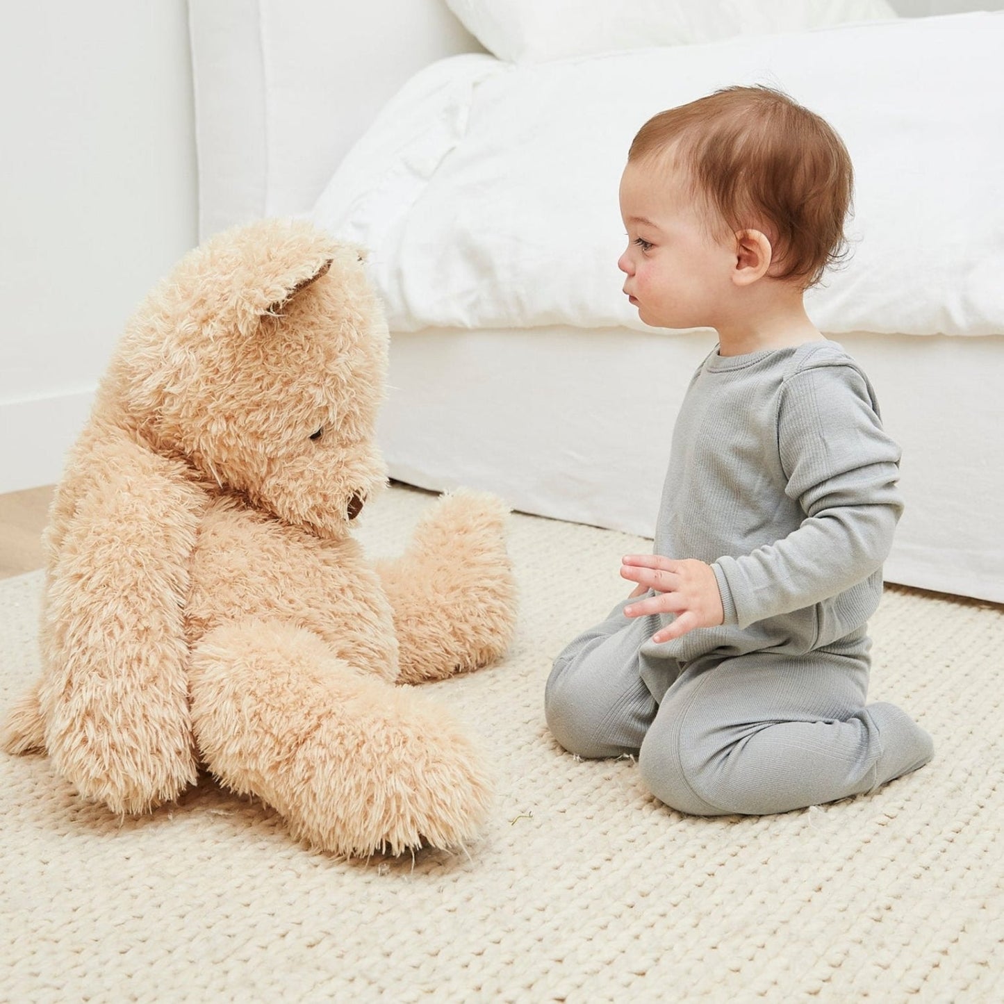 Baby in gray pajamas sitting next to a large teddy bear on a light-colored rug.