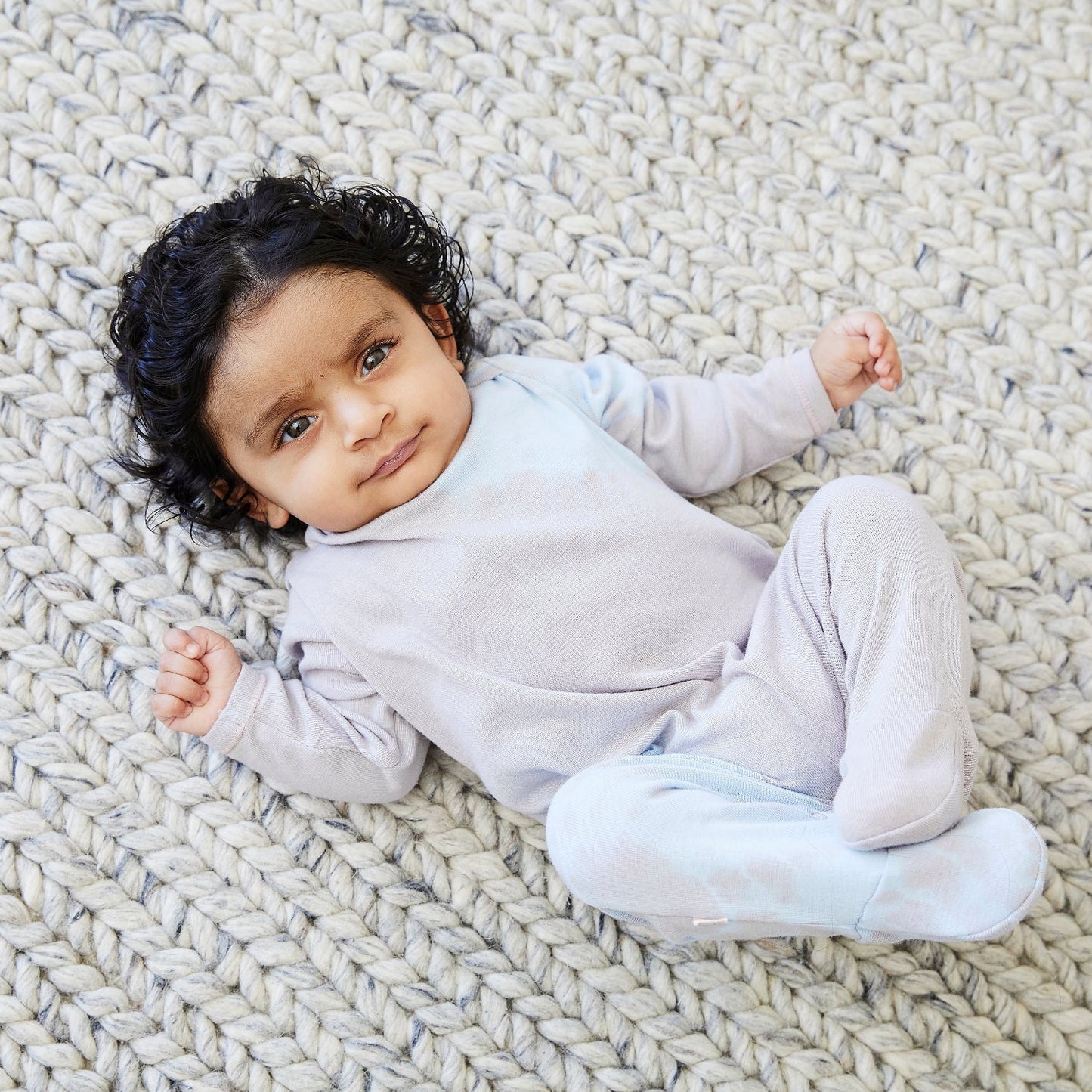 Baby lying on a textured rug wearing a light gray outfit
