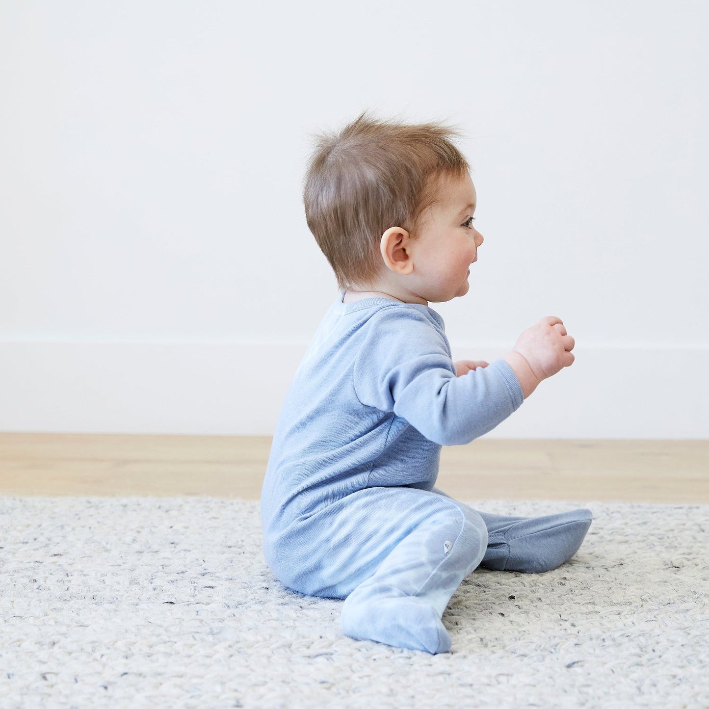 Baby in a blue onesie sitting on a textured rug with a plain background
