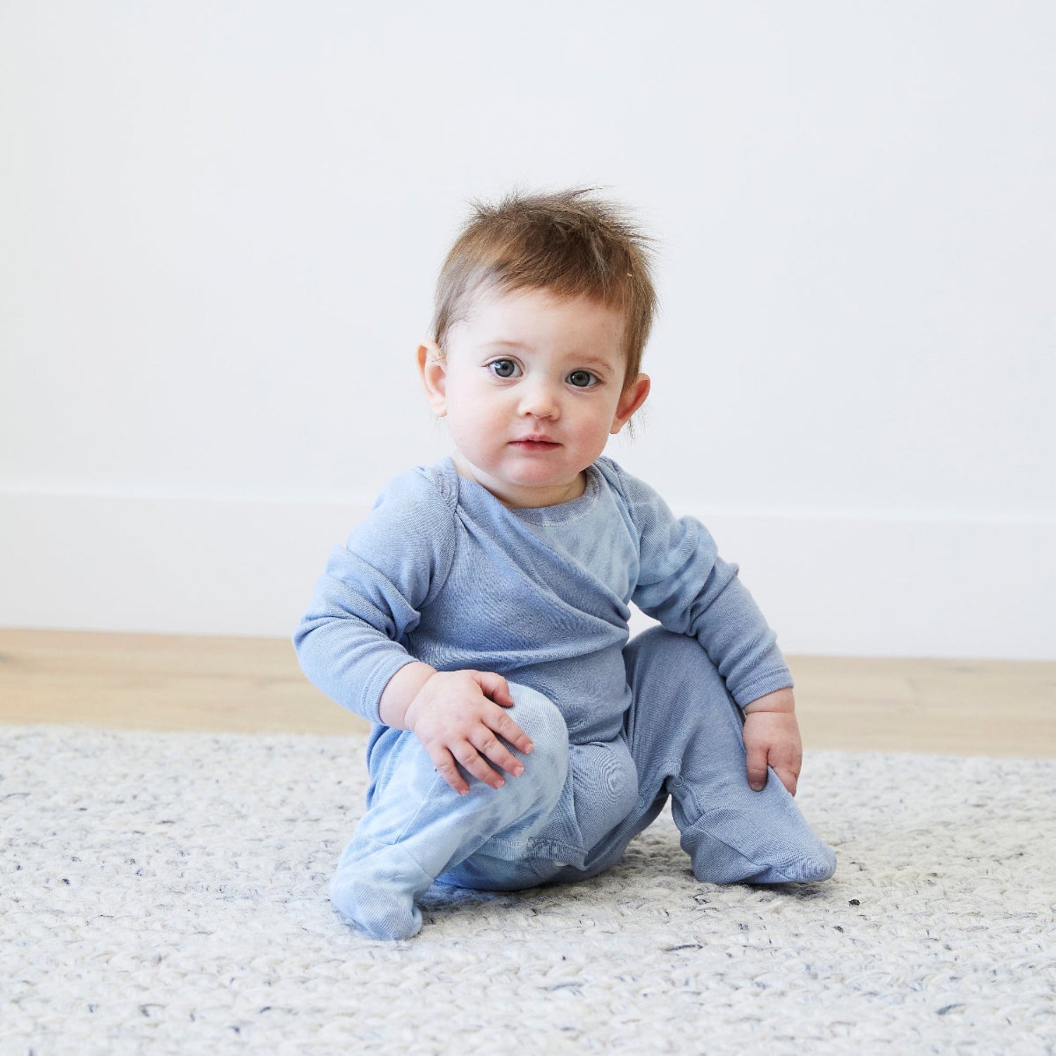 Baby wearing a light blue outfit sitting on a textured rug with a white background