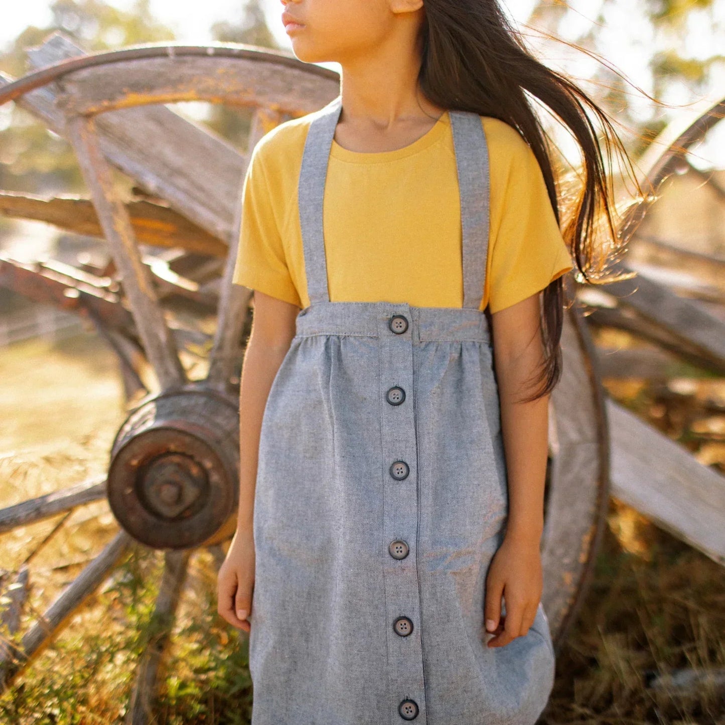 Person wearing a yellow shirt and blue denim dress with suspenders in front of a rustic wagon.