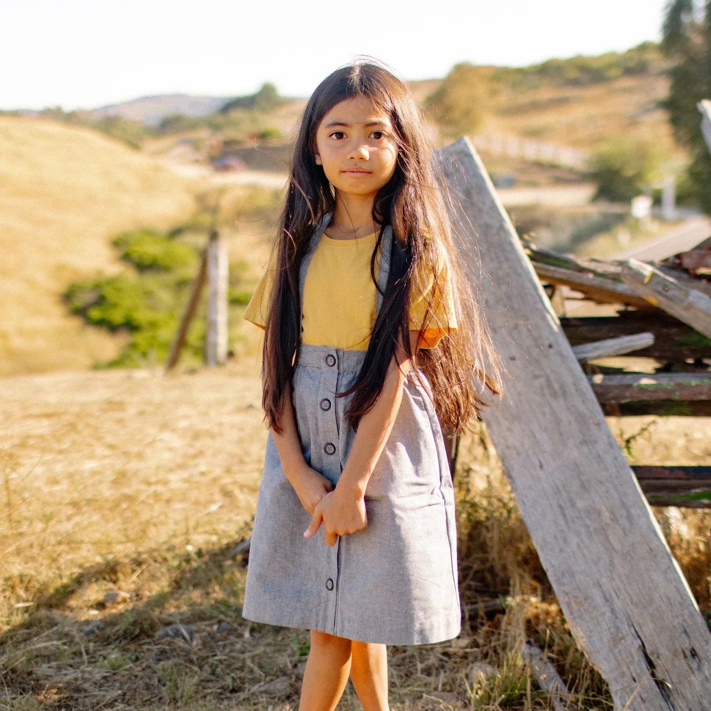 Young girl in a yellow top and gray skirt standing in a field with wooden planks and hills in the background.