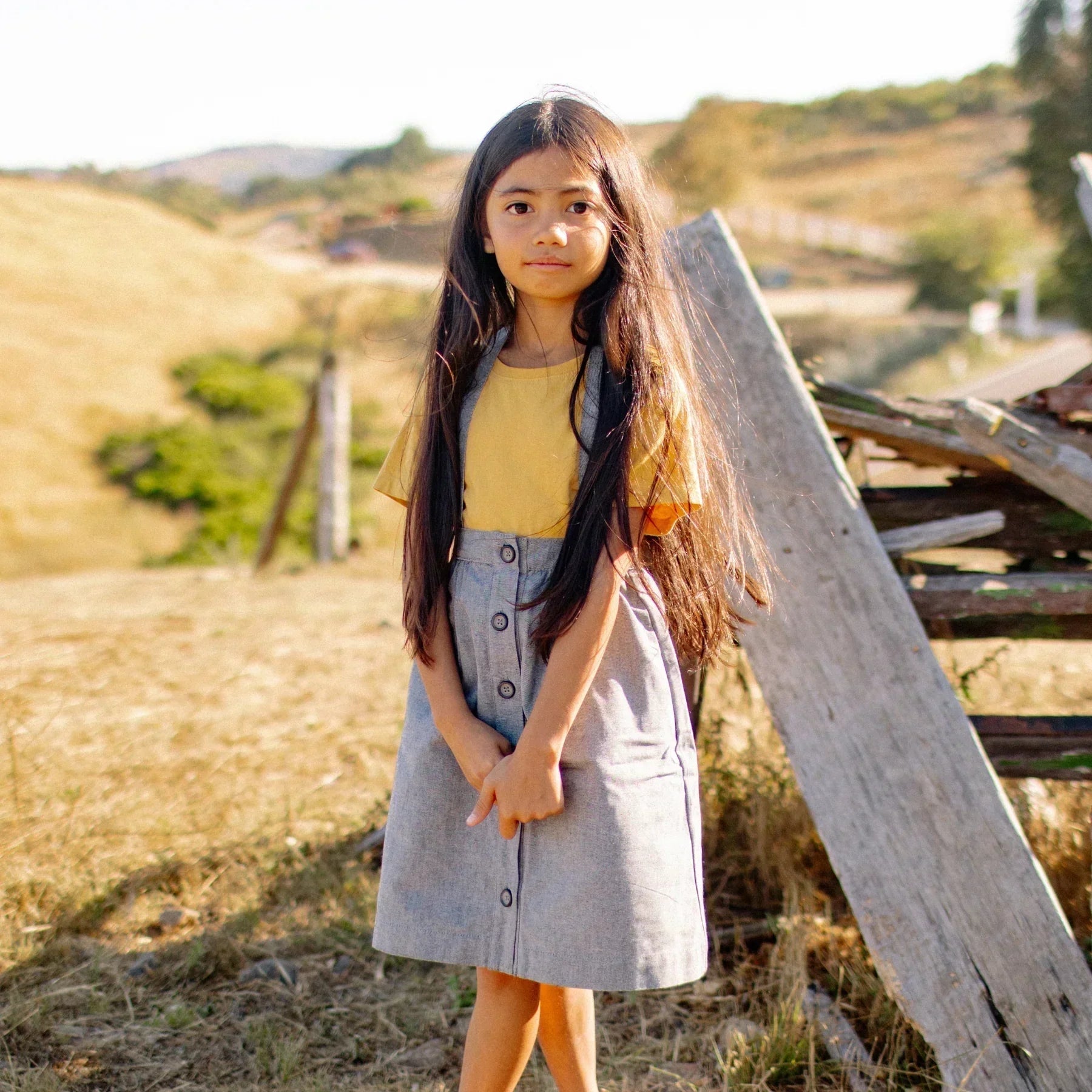 Young girl in a yellow top and gray skirt standing in a field with wooden planks and hills in the background.