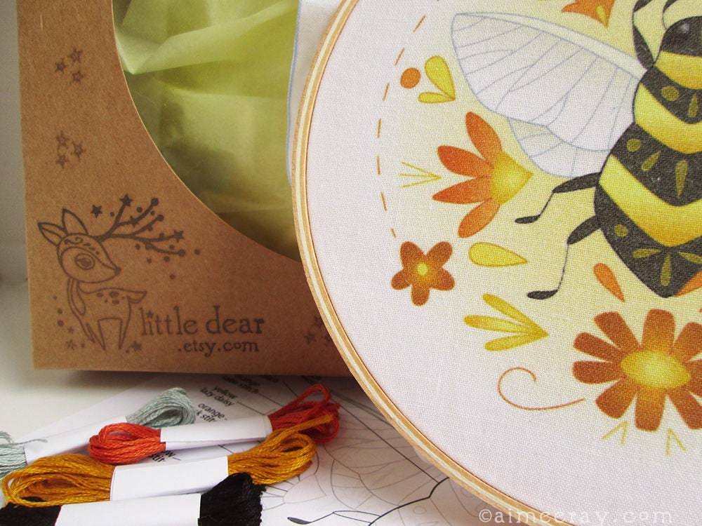 Embroidery hoop with bee and flower design, surrounded by embroidery threads and packaging.