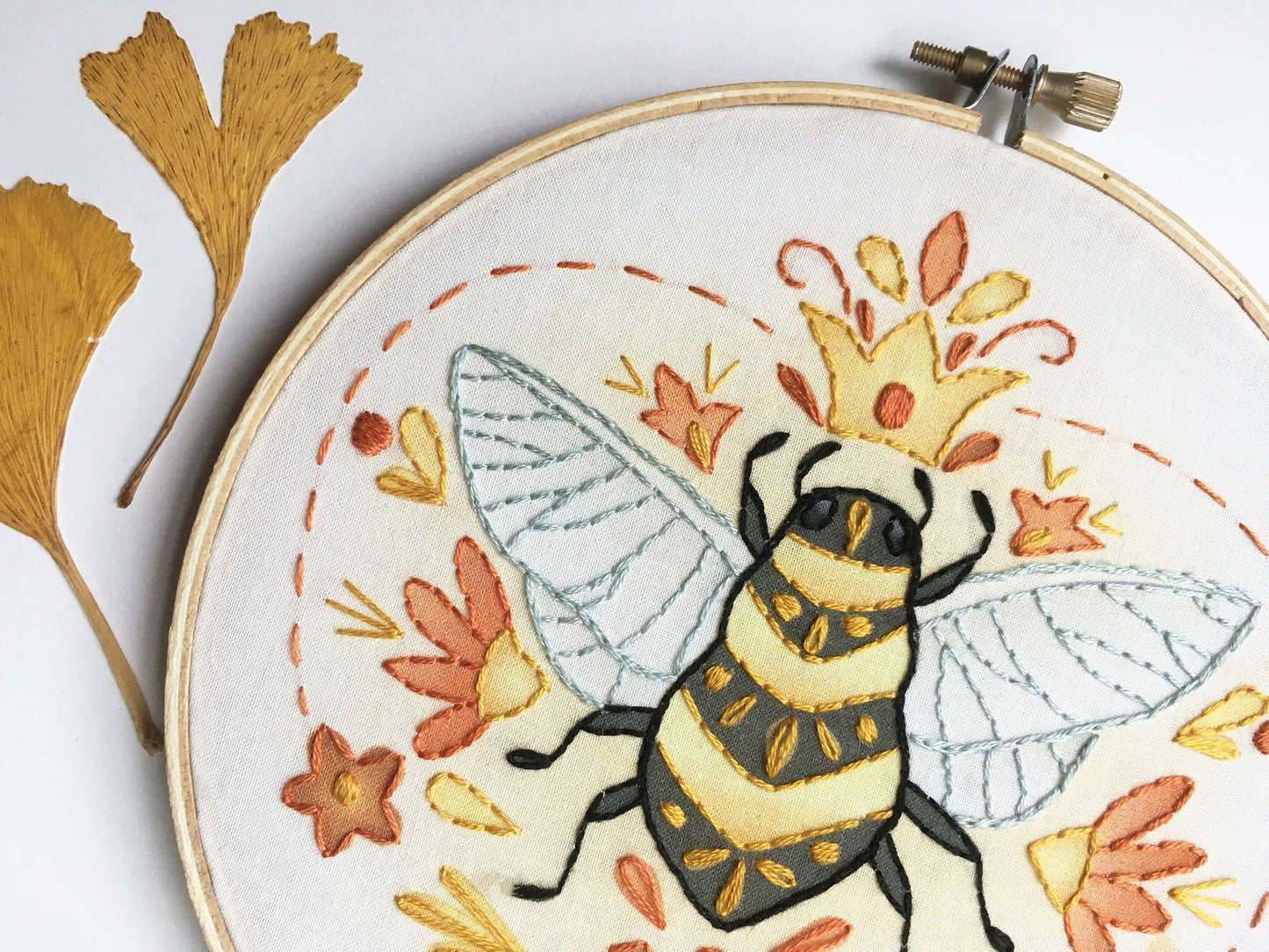 Embroidery hoop with a bee design surrounded by autumn leaves on a white background
