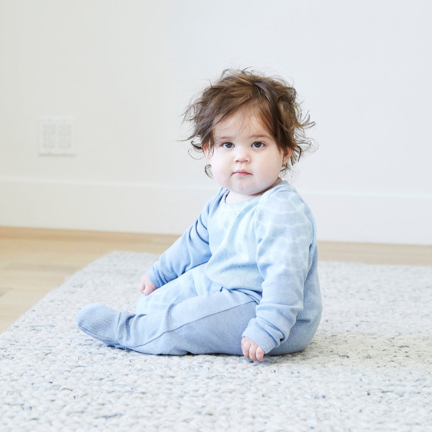 Baby in a light blue outfit sitting on a textured rug with a plain background