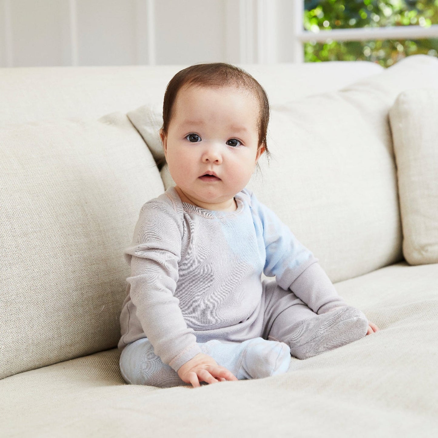 Baby sitting on a light-colored couch wearing a gray outfit.