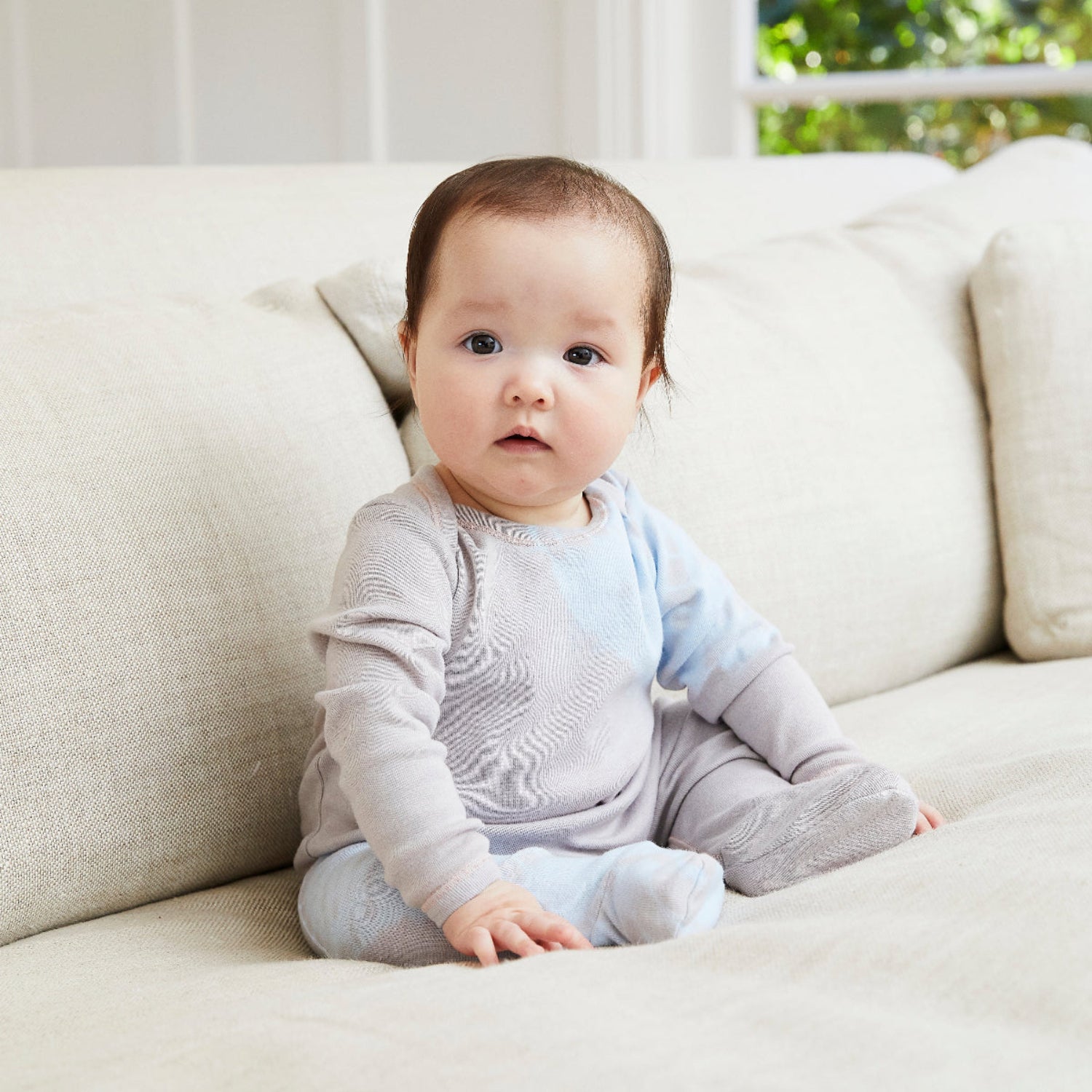 Baby sitting on a light-colored couch wearing a gray outfit.