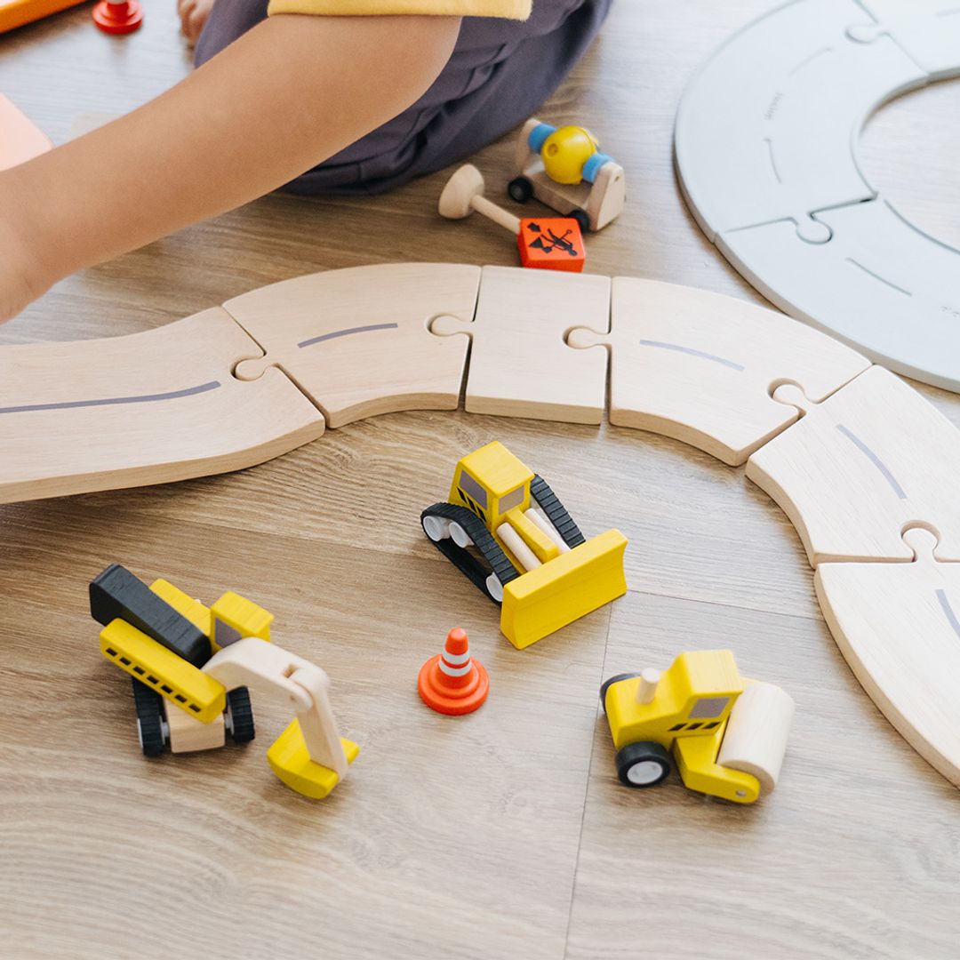 Toy construction vehicles on a wooden floor with a child's hand and puzzle in the background.