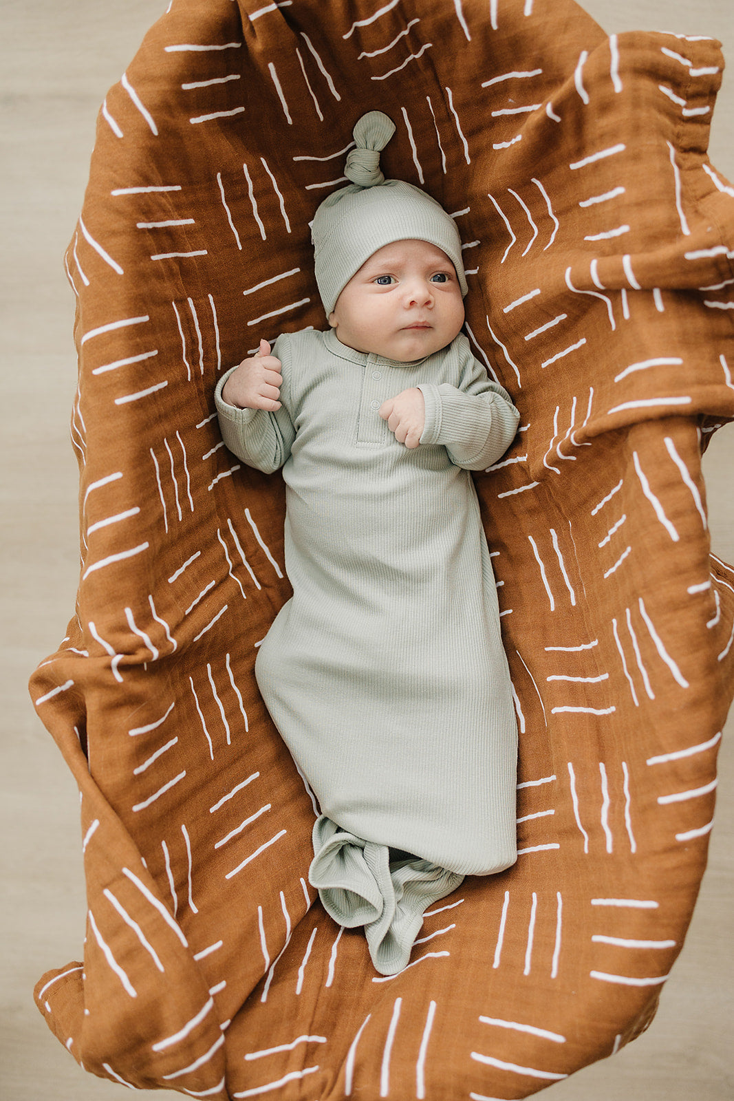 Baby wrapped in a light green swaddle and cap, lying on a brown blanket with white patterns.