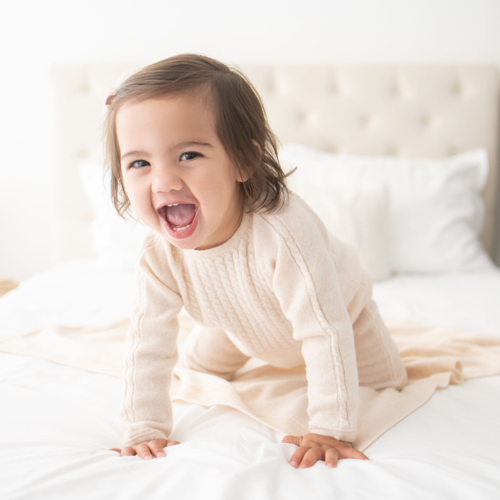 baby - toddler  in a white sweater sitting on a bed with a happy expression
