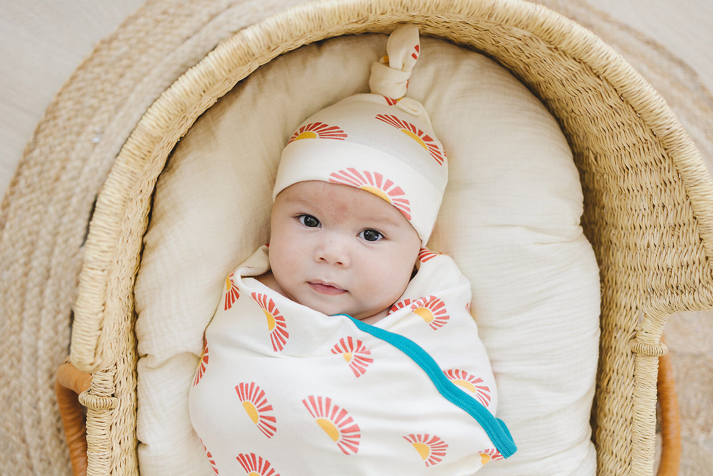 Baby swaddled in a patterned blanket and wearing a hat, lying in a moses basket.