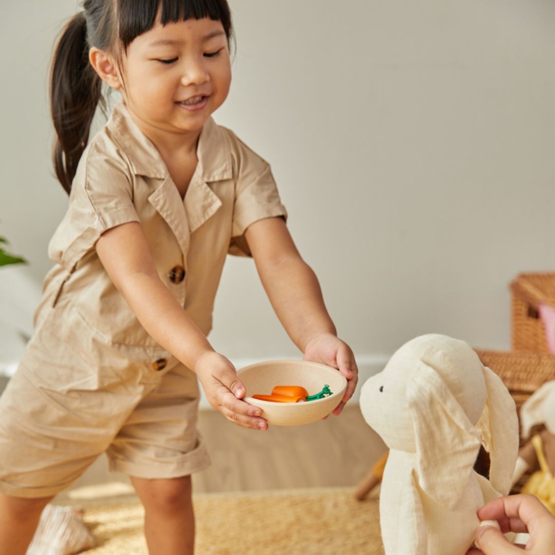 Child in a beige outfit holding a bowl with toys, interacting with a plush toy in a home setting.