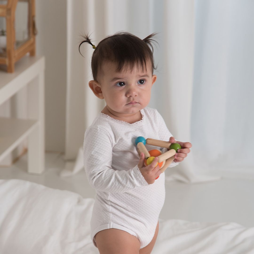 Baby in a white onesie holding wooden toys in a softly lit room.