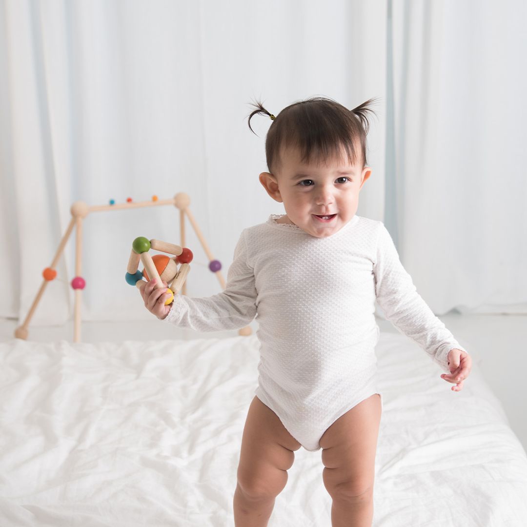 Baby in a white onesie standing on a white surface with a colorful baby gym in the background.