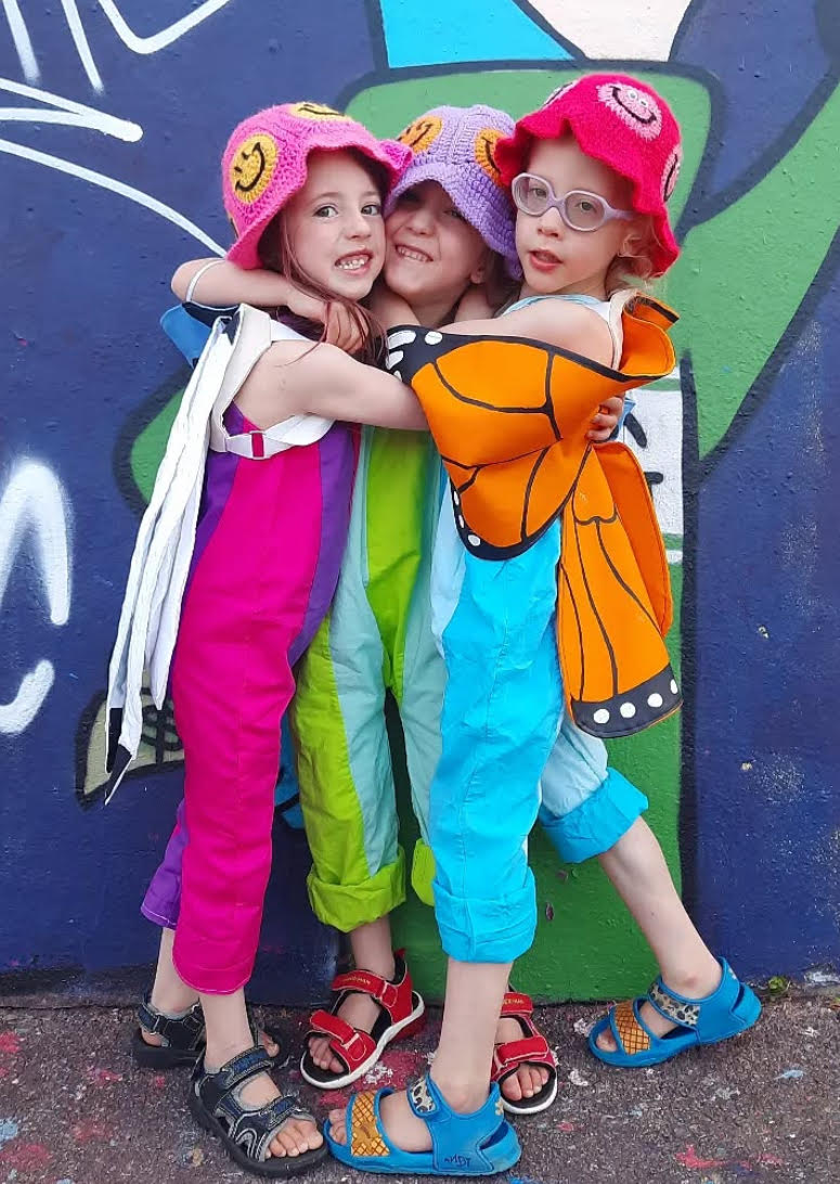 Three children in colorful costumes posing against a graffiti-covered wall.