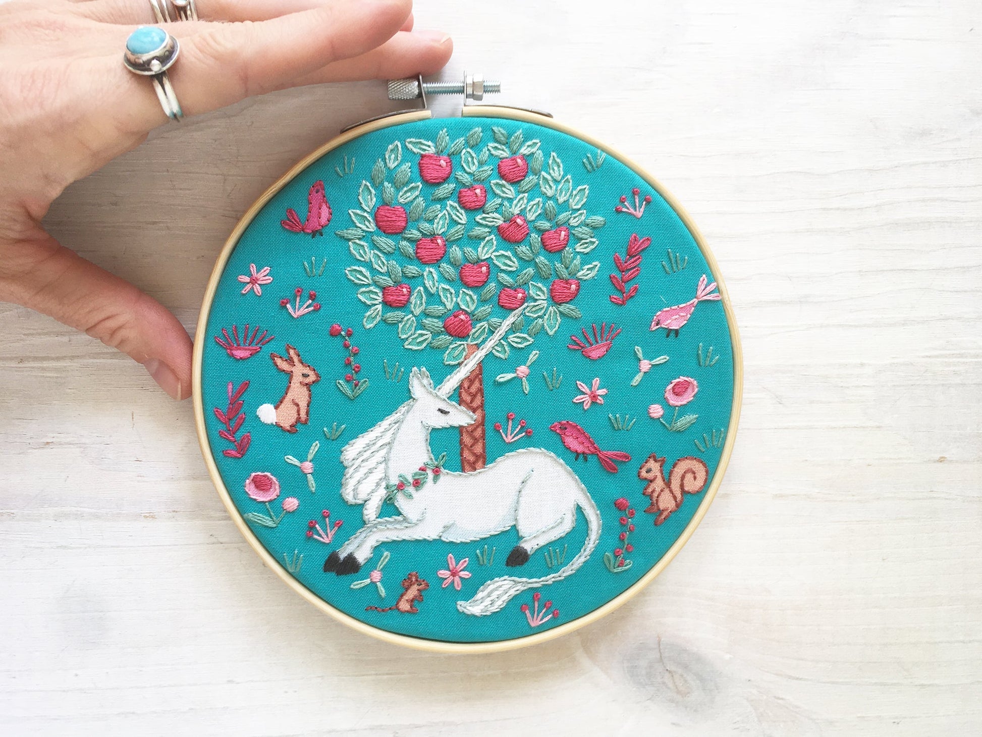 Embroidery hoop with a unicorn design on a teal background, held by a hand.