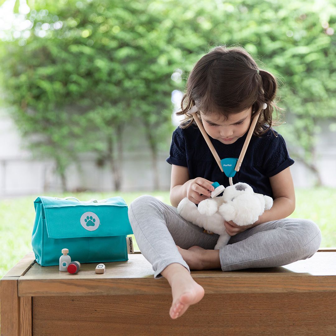 Child playing with a plush toy outdoors, with a teal bag and items on a table in front.