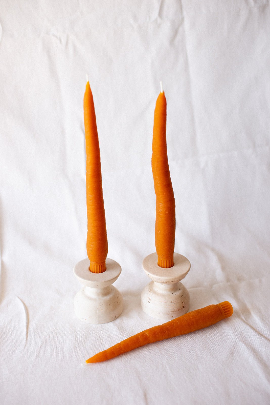 Two tall orange candles in white candle holders with a carrot on a white background