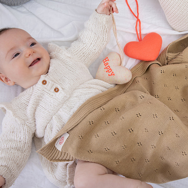 Baby wearing a white knit outfit with a brown textured blanket and heart-shaped toys.