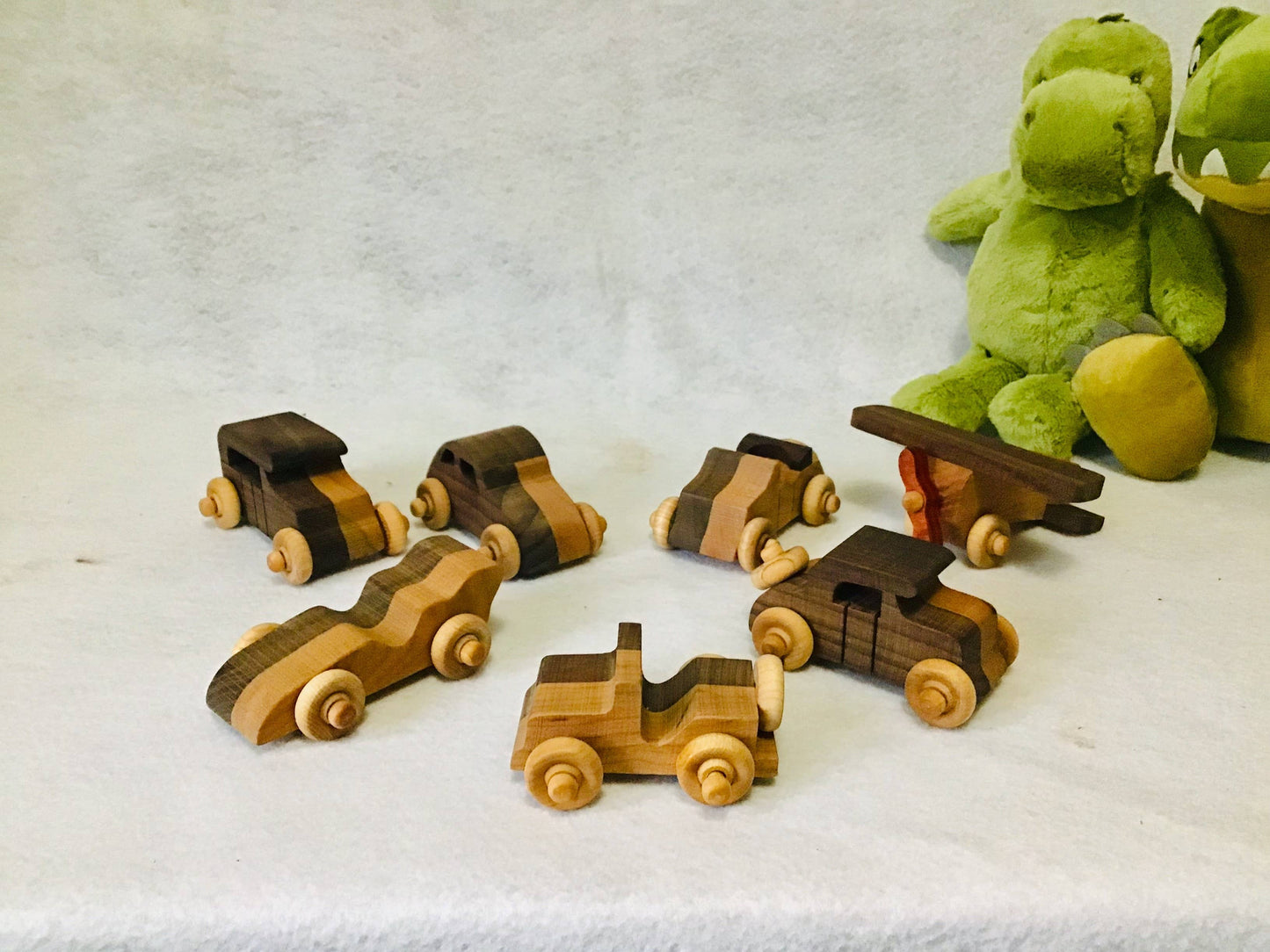 Set of wooden toy vehicles on a white surface with a green stuffed dinosaur in the background.