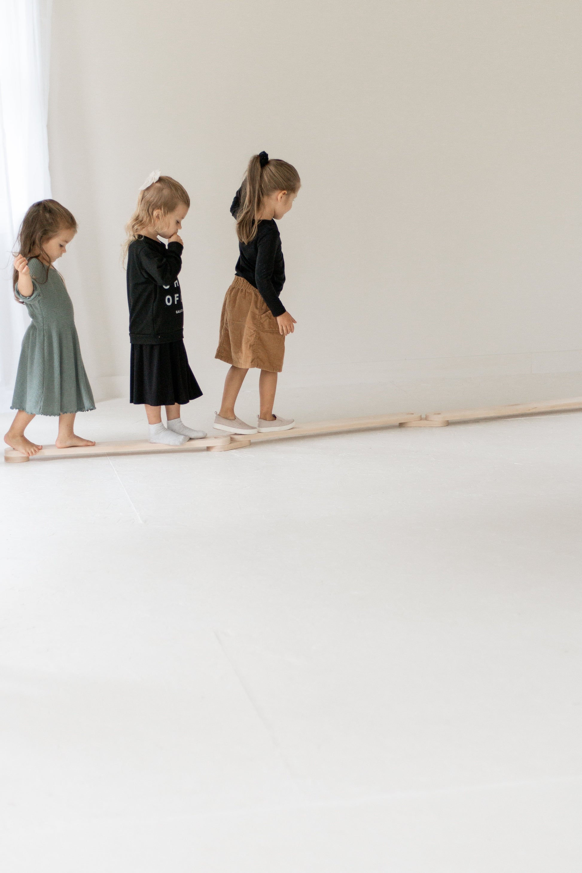 Three children standing on a wooden balance beam against a white wall.