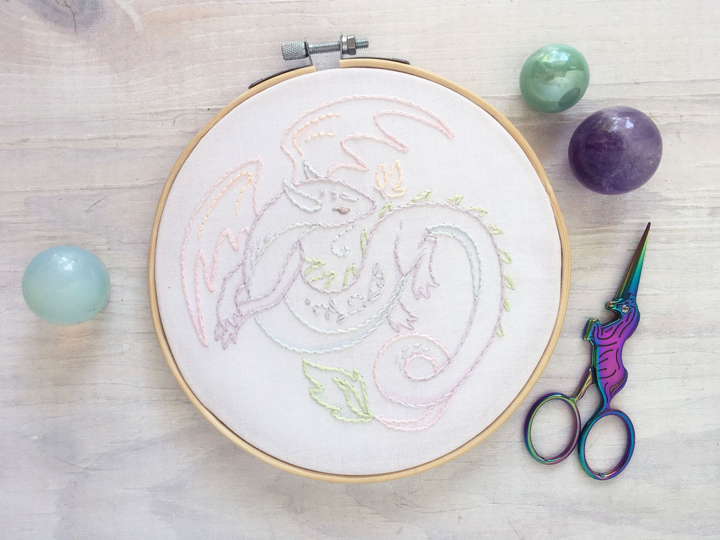 Embroidery hoop with colorful dragon design on a wooden surface with scissors and stones.