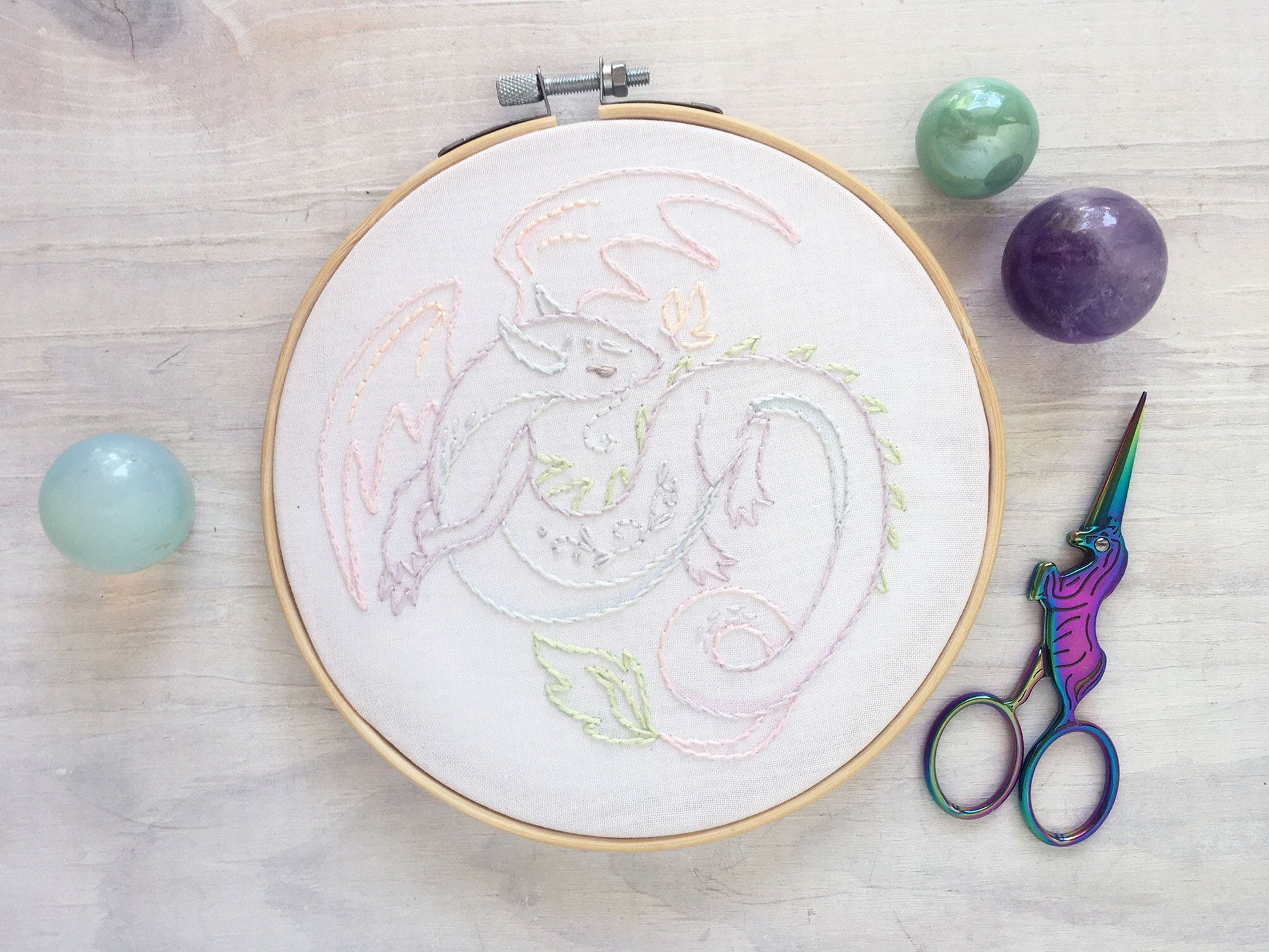 Embroidery hoop with colorful dragon design on a wooden surface with scissors and stones.