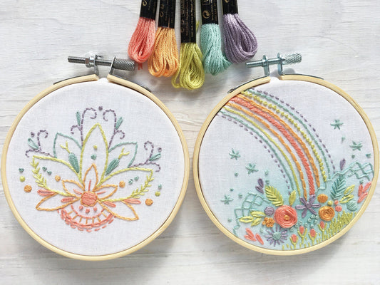 Two embroidery hoops with floral and rainbow designs, accompanied by colorful thread spools on a light wooden surface.