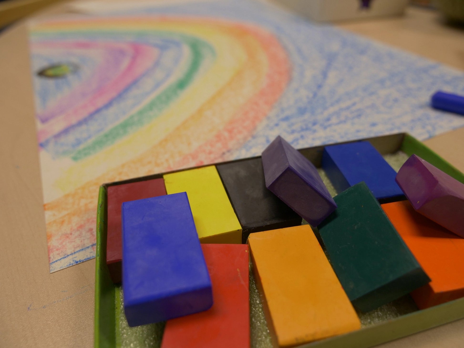 Colorful crayon blocks in a box in front of a child's drawing of a rainbow