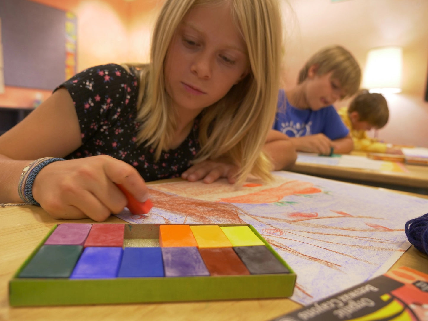 Child using colorful block crayons to draw on a table with other children in the background