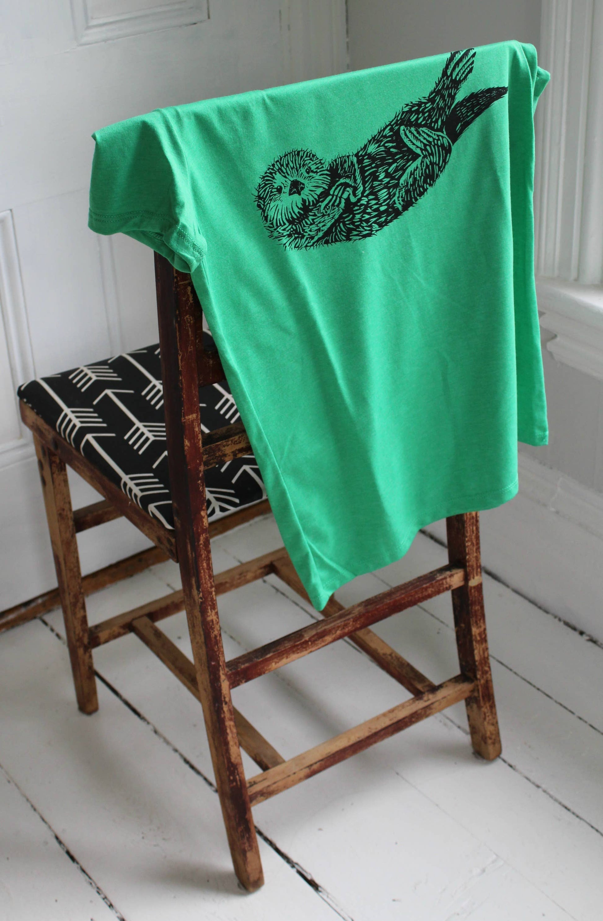 Green t-shirt with a black otter design draped over a wooden chair.