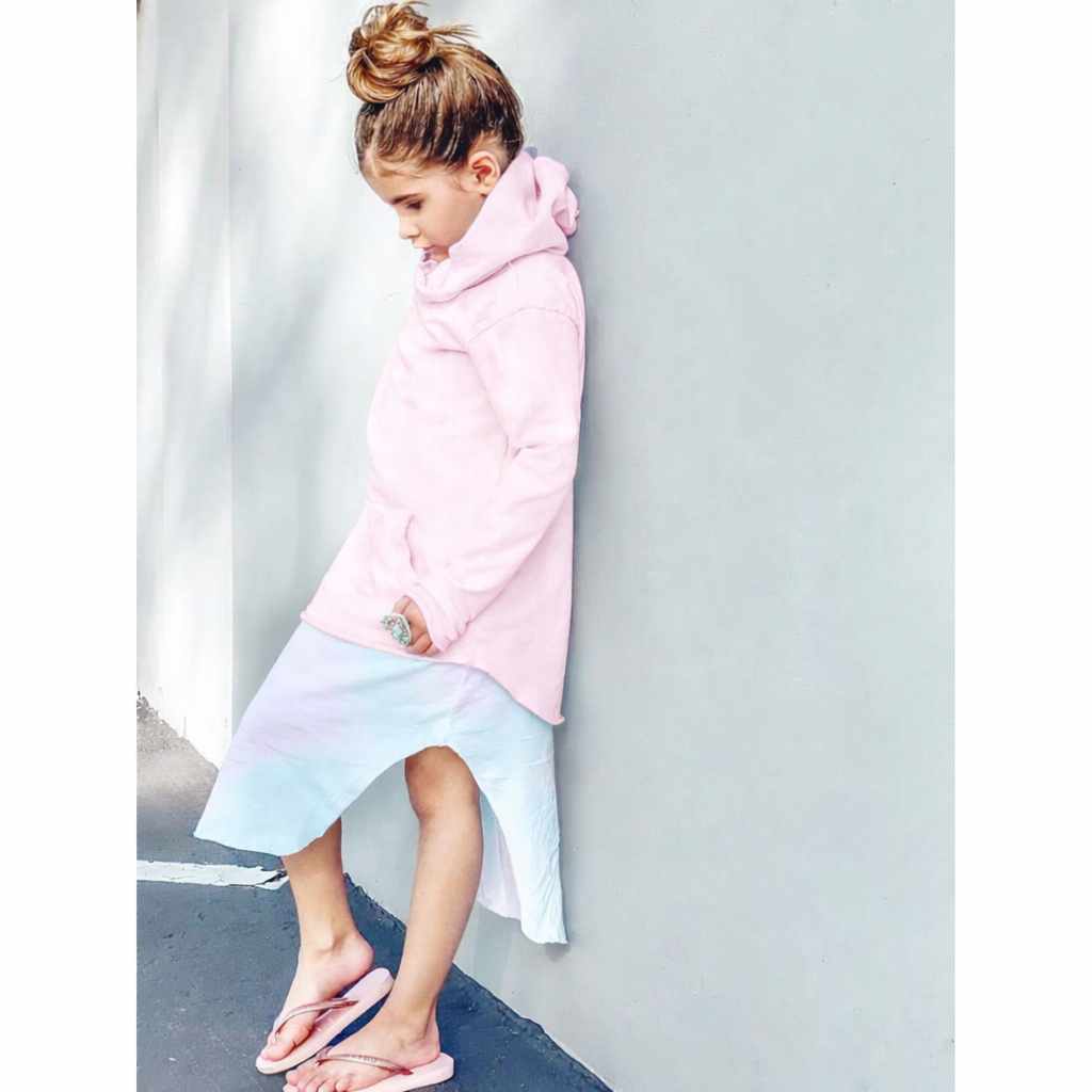 A young girl with her hair in a bun leans against a light gray wall, wearing a light pink hooded sweatshirt and a pastel tie-dye skirt with pink flip-flops.