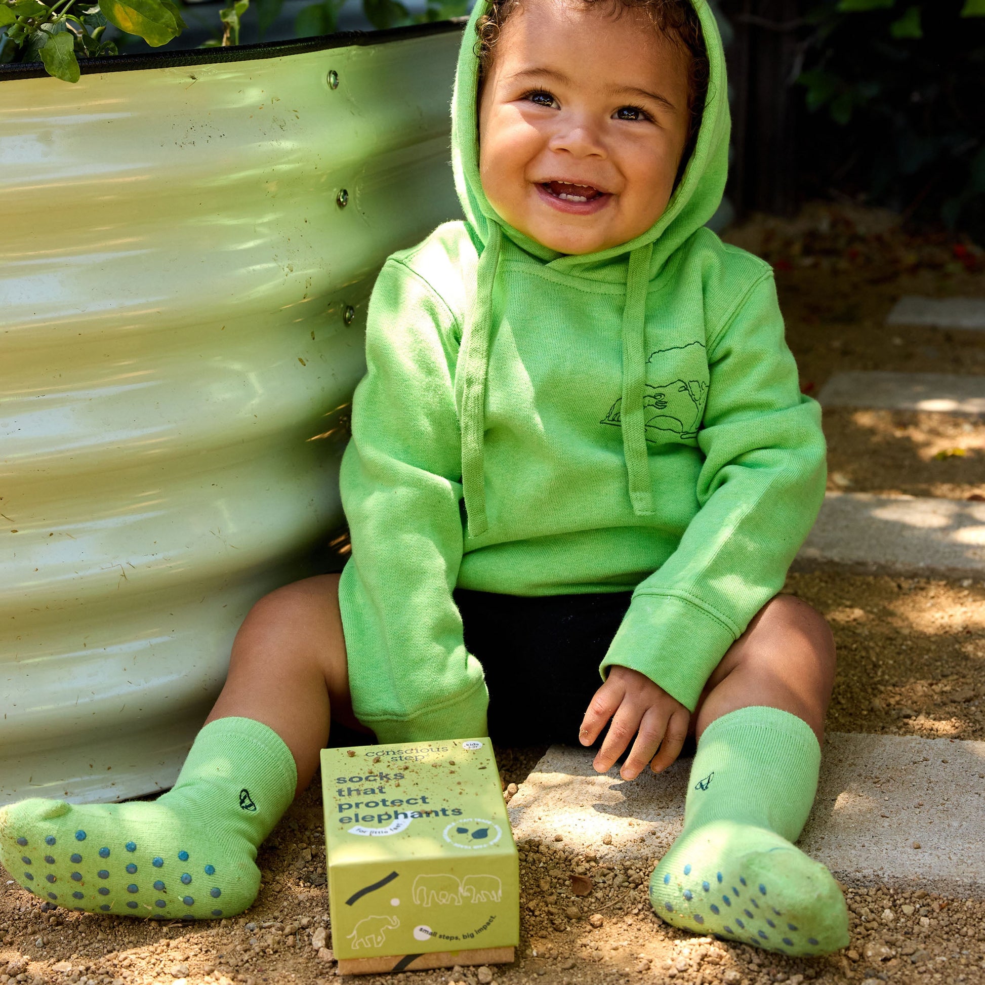 Child wearing a green hoodie and matching shoes sitting next to a box with a plant in the background