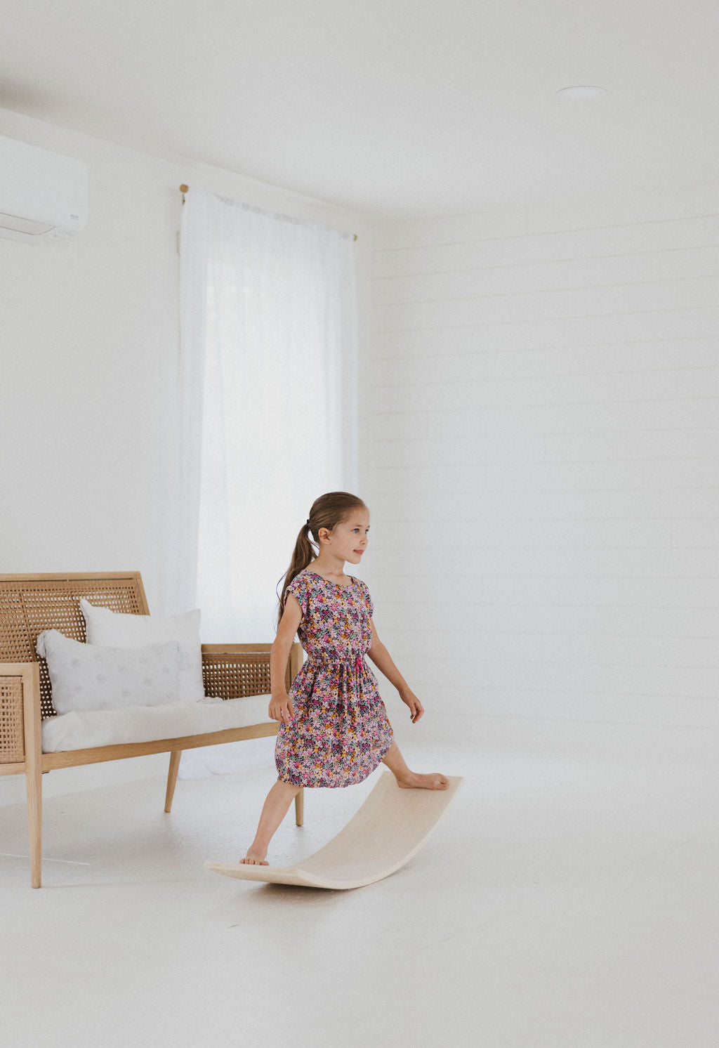 Young girl in a floral dress standing on a white rug in a minimalistic room with a wooden bench and white walls.
