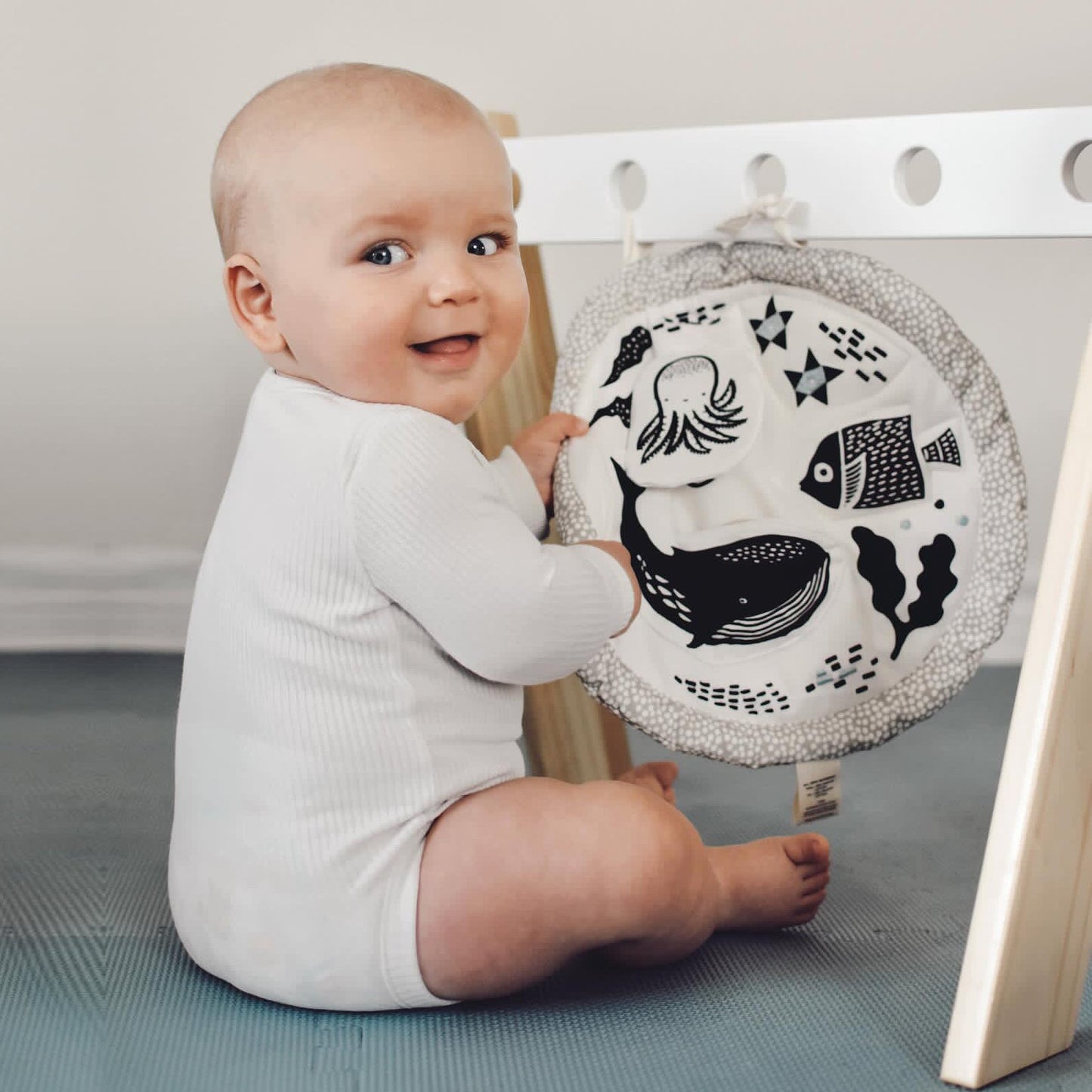 Baby holding a round wooden toy with black illustrations on a blue mat.