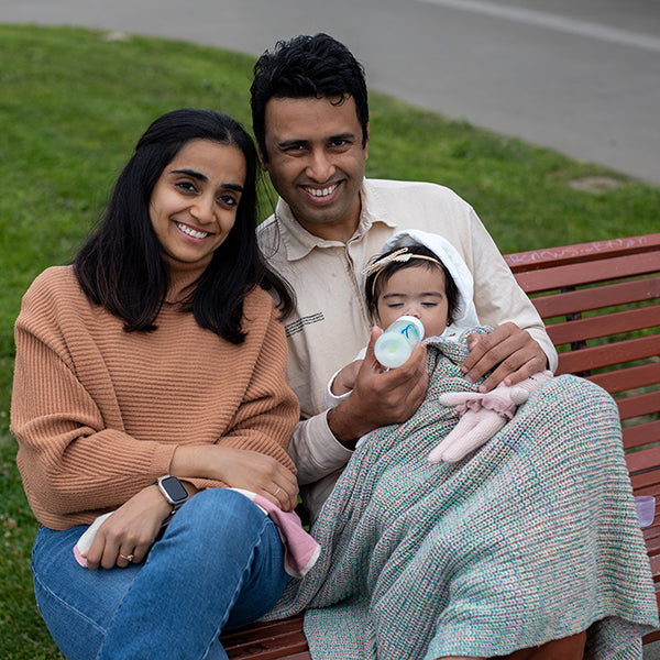 Family of three sitting on a park bench with a baby wrapped in a blanket.