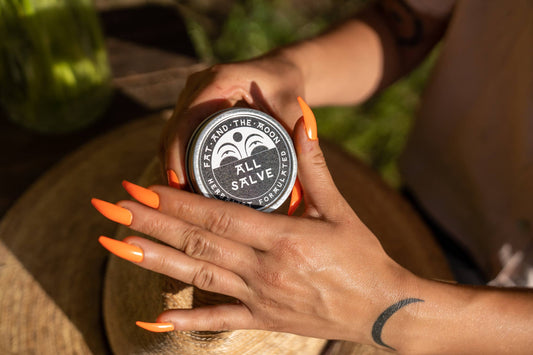 Person with bright long orange nails holding a jar of 'All Salve' with a brand logo, surrounded by greenery.
