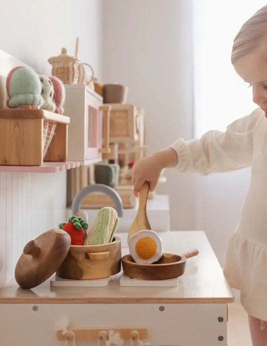a child playing with wooden pots and pans toys and crochet foods in a play kitchen