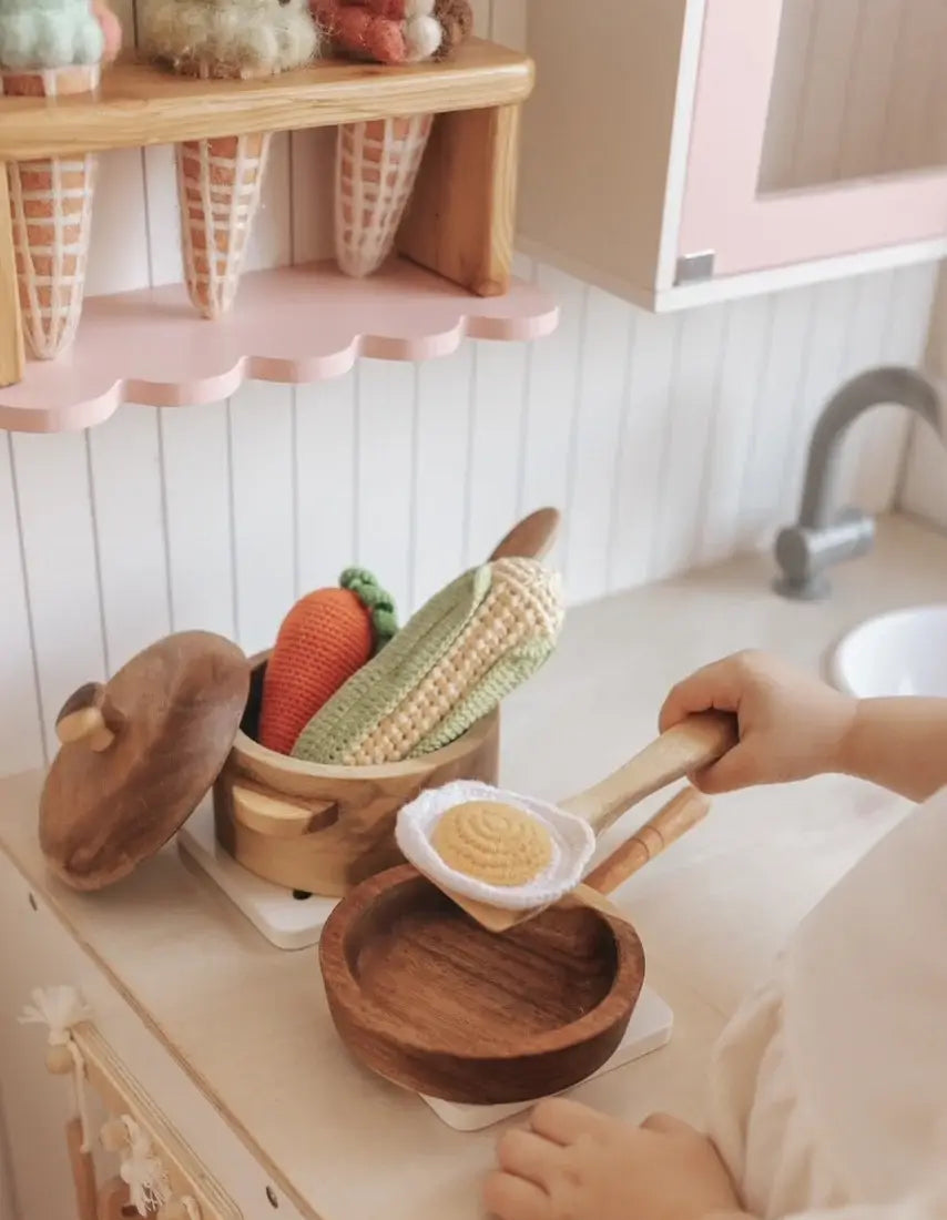 a child playing in a play kitchen with wooden pots and pans, picking up a crochet egg with a wooden toy spoon