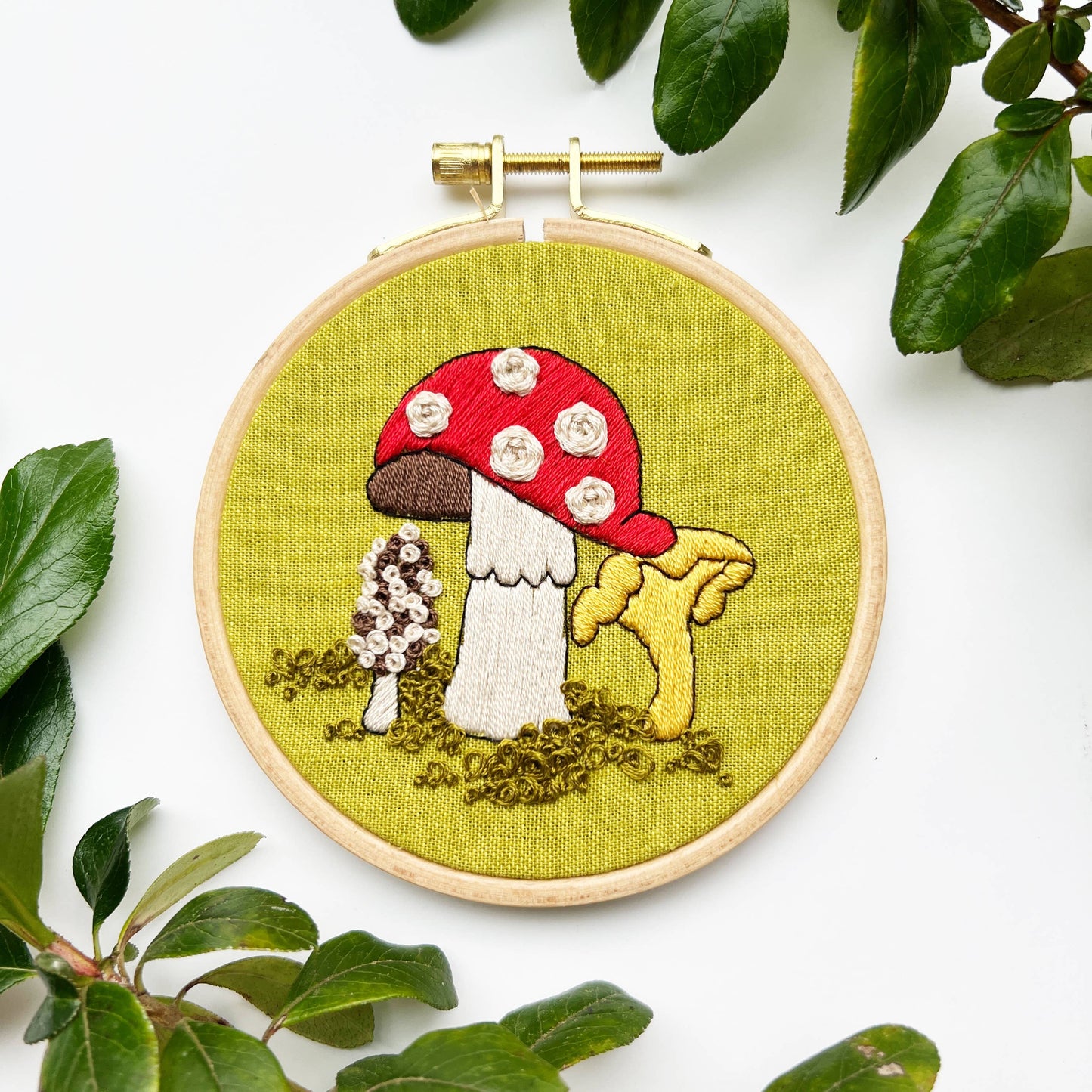 Embroidery hoop with a mushroom design surrounded by green leaves on a white background