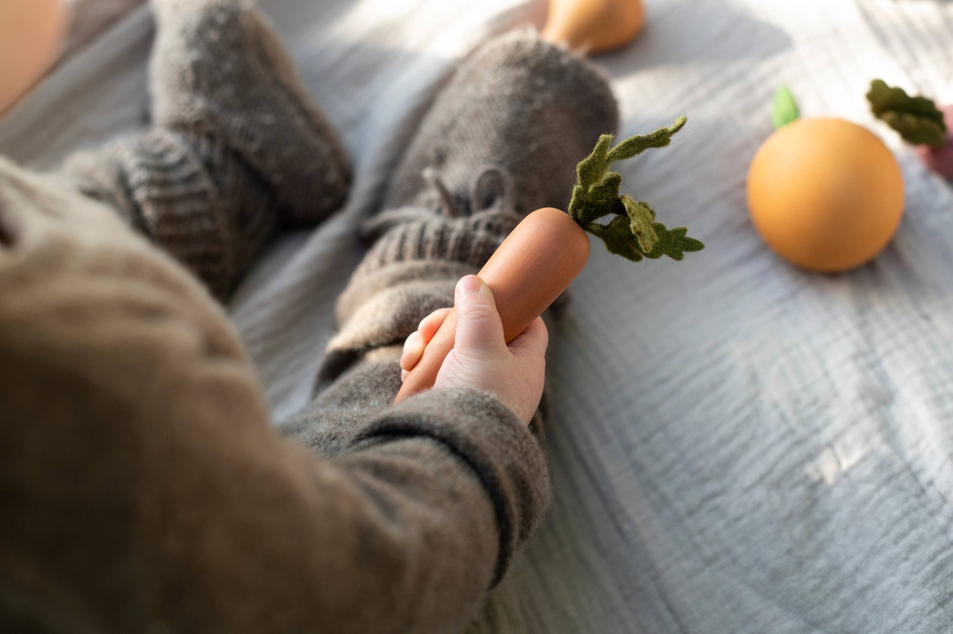 a child sitting on a bed holding a wooden toy carrot with more wooden fruits and vegetabes next to them