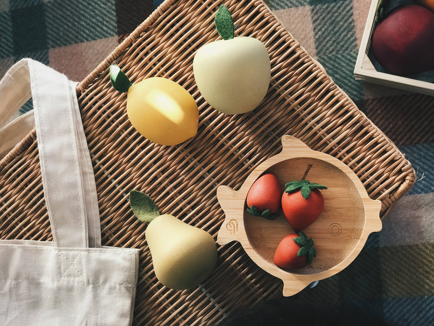  wooden pear, lemon, and apple on a wicker basket next to a cotton tote bag and three wooden strawberries in a fish-shaped bowl