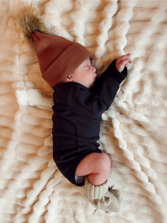 Baby wearing a brown knit hat and black onesie lying on a textured cream blanket.