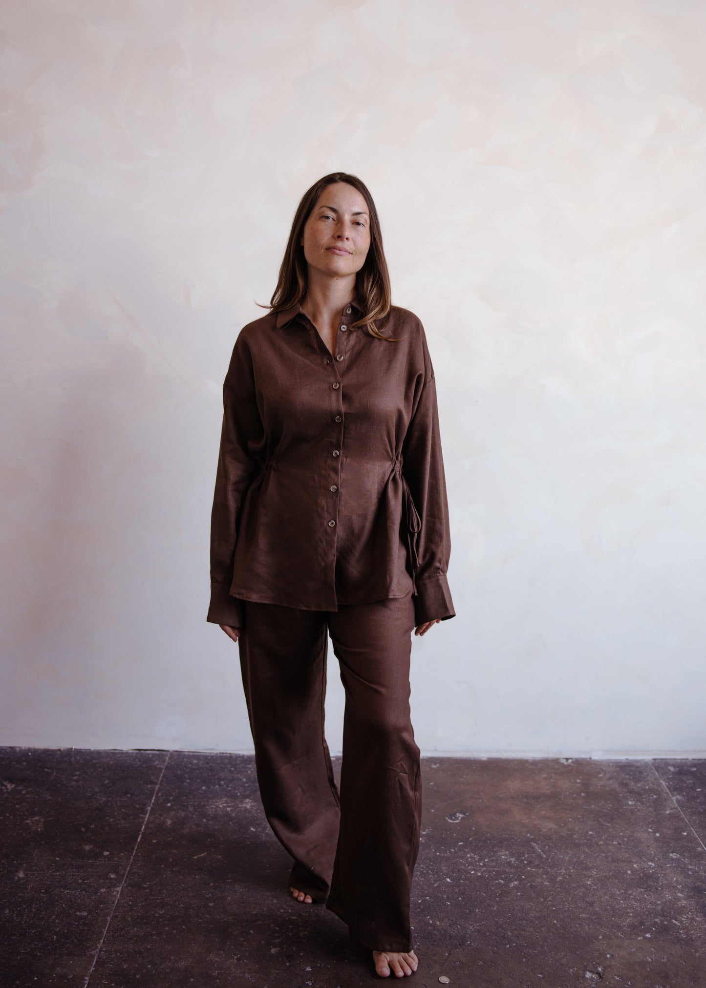 Woman wearing a brown shirt and pants standing against a plain wall.