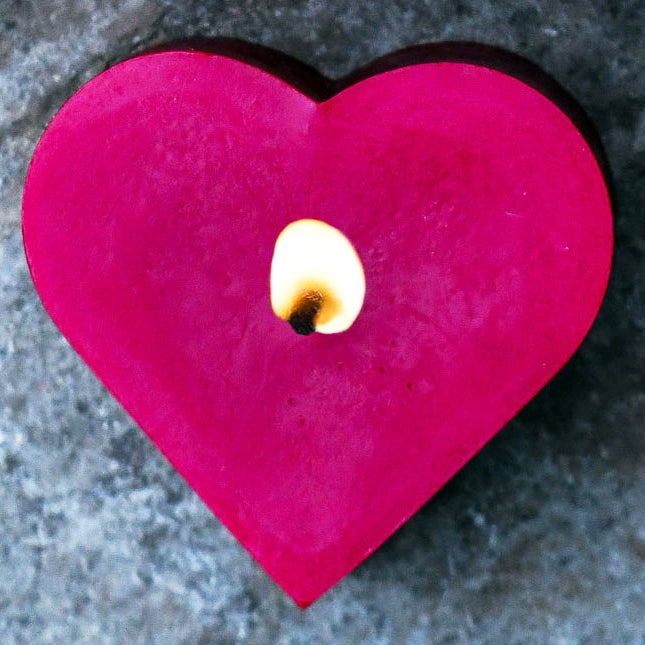 A red heart-shaped beeswax candle with a lit flame in the center, placed on a dark surface.